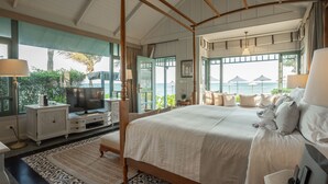 Una cama con dosel y vistas al océano, un televisor sobre un mueble blanco y un ventanal con cortinas.