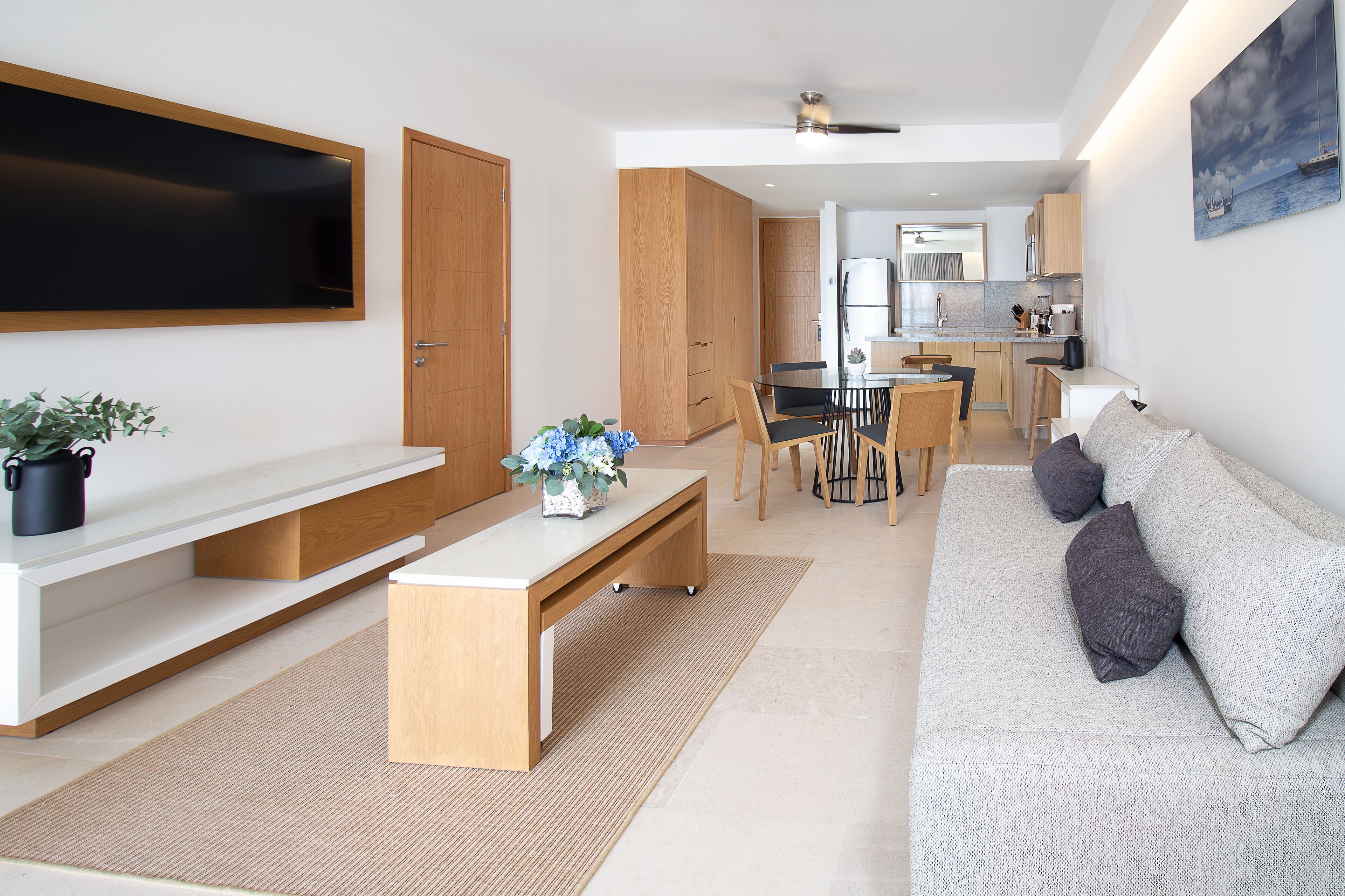 Una sala moderna con un televisor de pantalla plana, un banco de madera, un sofá con cojines y un comedor con sillas y una mesa.