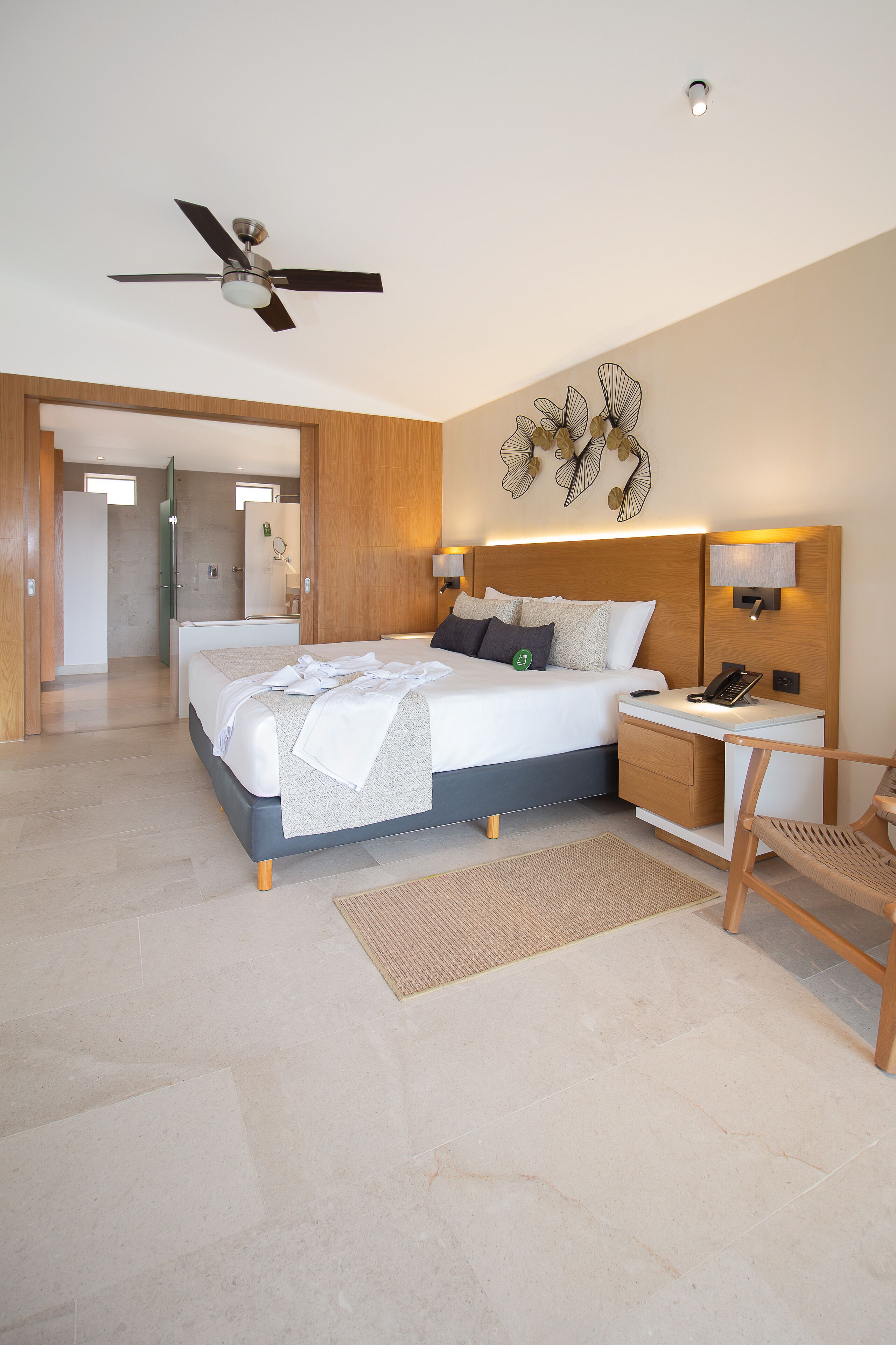 Una camera d'albergo moderna con un letto grande, un comodino, una scrivania, una sedia, una ventola a soffitto e un bagno visibile sullo sfondo.