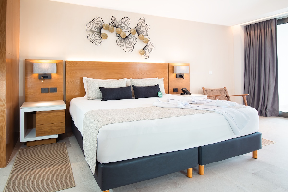A modern hotel room with a large bed, wooden headboard, bedside tables with lamps, and a decorative wall piece.