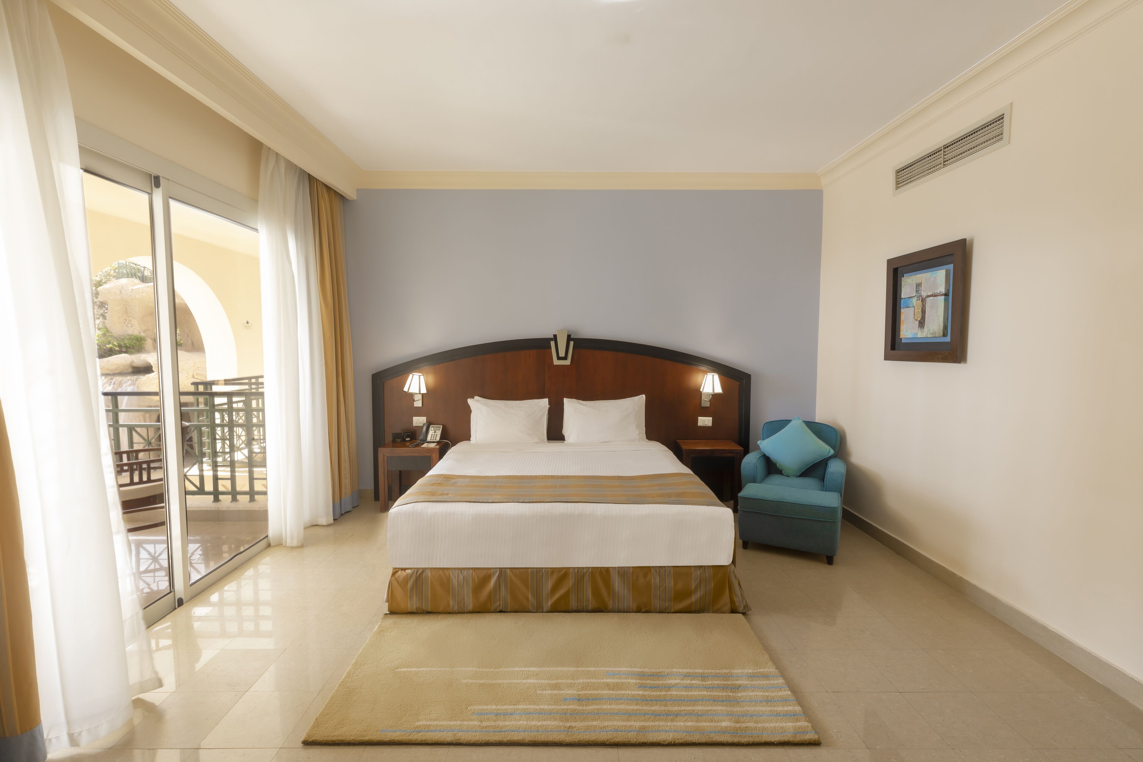 A hotel room with a large bed, a small sofa, a balcony with outdoor furniture, and a painting on the wall.