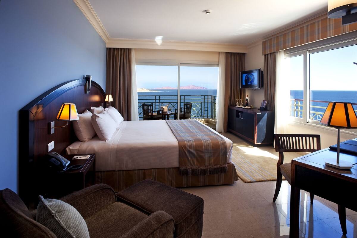 Habitación de hotel con una cama grande, un escritorio, un televisor y un balcón con vista al mar.