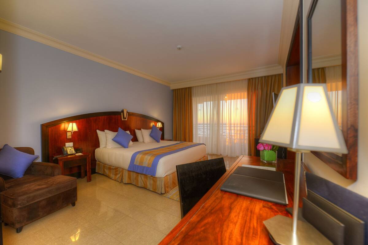 Una habitación de hotel con cama, escritorio, silla, lámpara y una mesita con un florero.