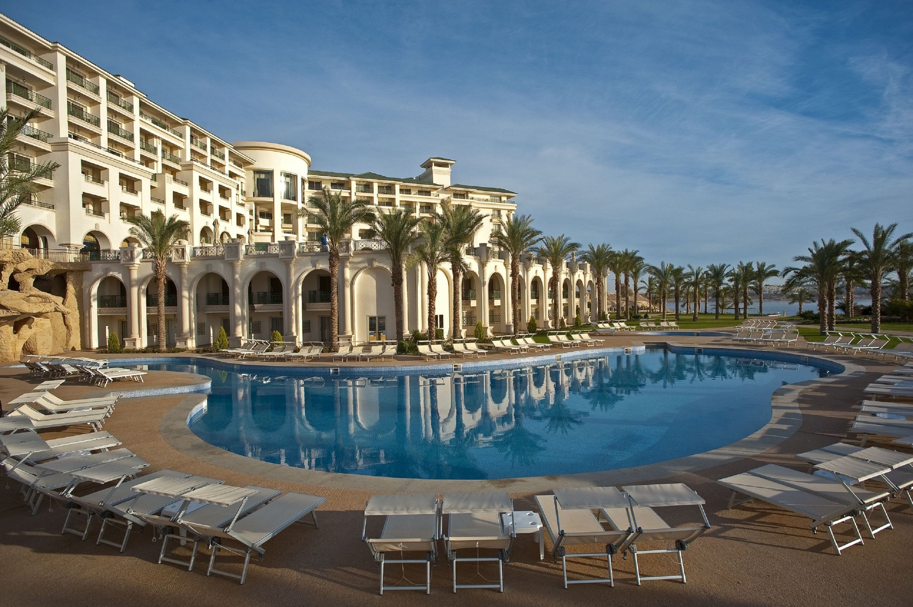 Ein Resort mit großem Pool, Liegestühlen, Palmen und einem mehrstöckigen Gebäude mit Balkonen.