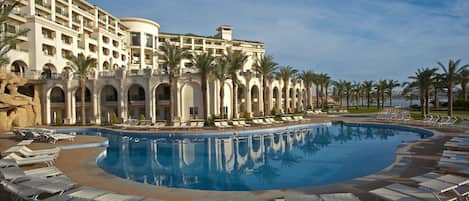 Ein Resort mit großem Pool, Liegestühlen, Palmen und einem mehrstöckigen Gebäude mit Balkonen.