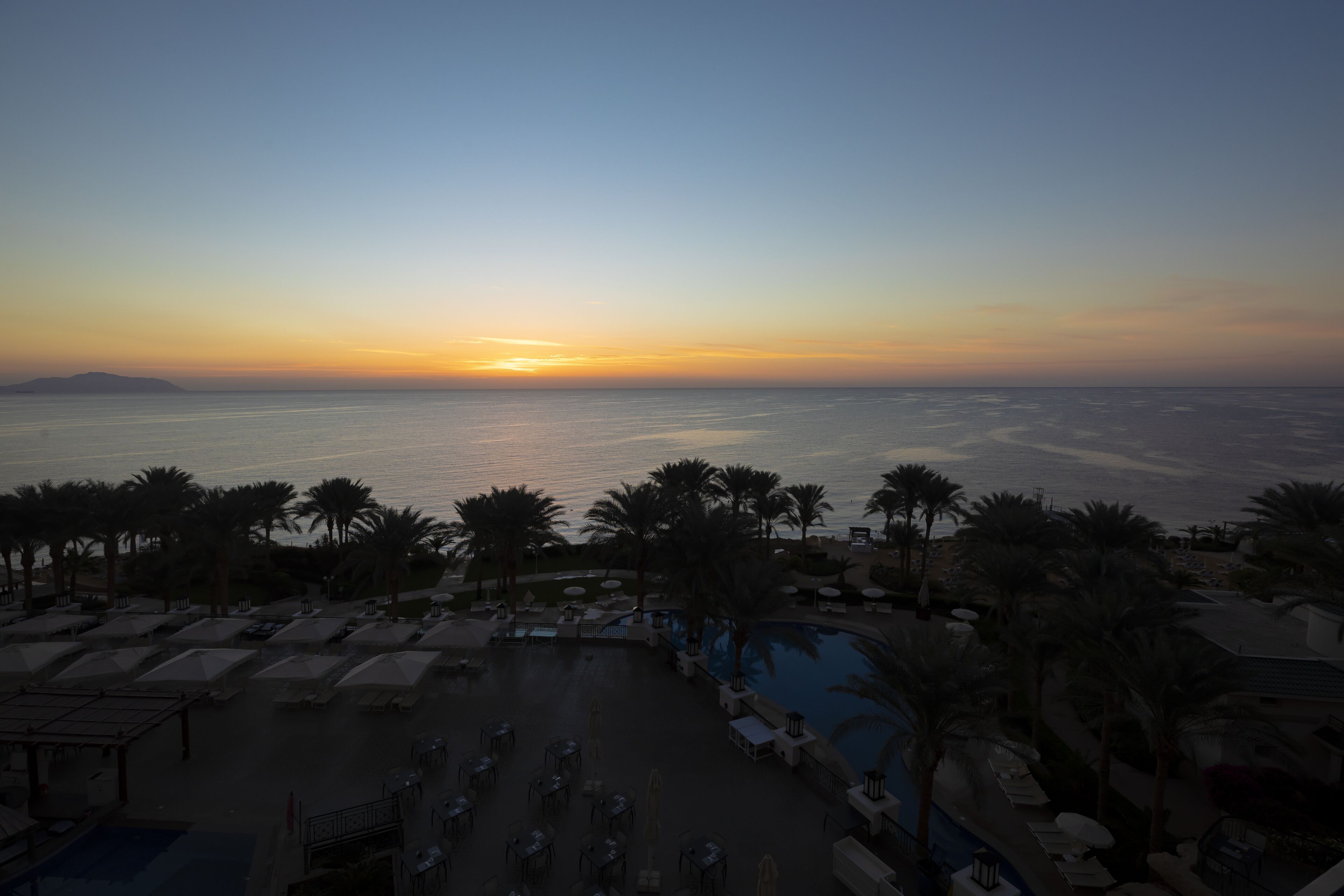 A resort pool area with sun loungers and umbrellas, a swimming pool, and a view of the ocean at sunset.