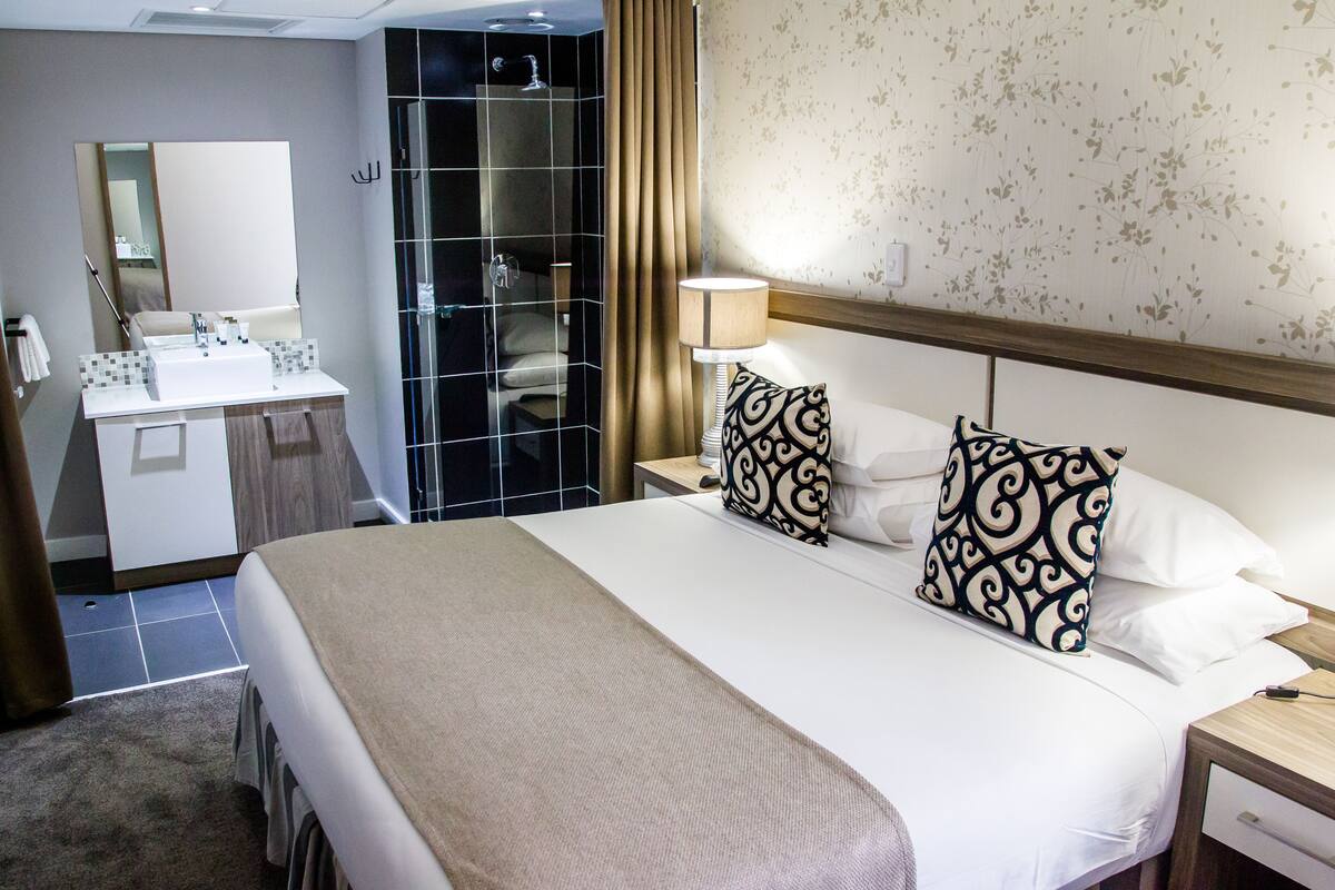 A hotel room with a bed, a bedside table, a nightstand, a mirror, a bathroom with a shower, and a vanity with a mirror and toiletries.