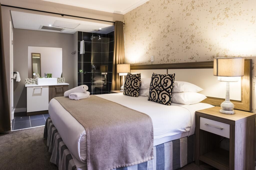 Una habitación de hotel con cama, mesita de noche, lámpara, baño con ducha y espejo.