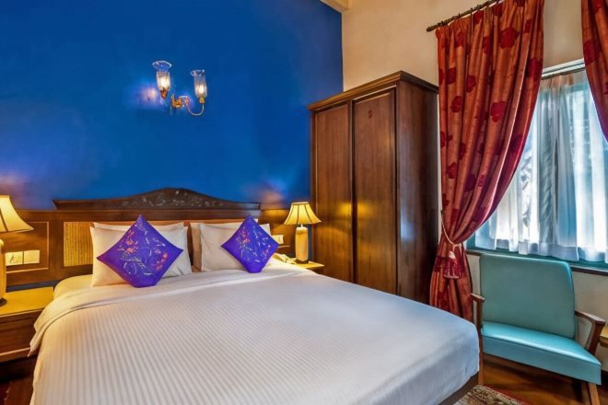 Une chambre d’hôtel avec un grand lit, deux lampes de chevet, une table de nuit, une armoire en bois et une fenêtre avec des rideaux rouges.