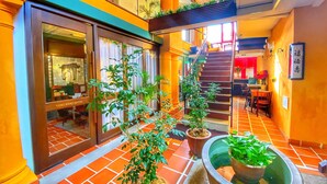 Un pasillo de hotel con suelo de baldosas, plantas en macetas, una escalera y un comedor con sillas y una mesa.