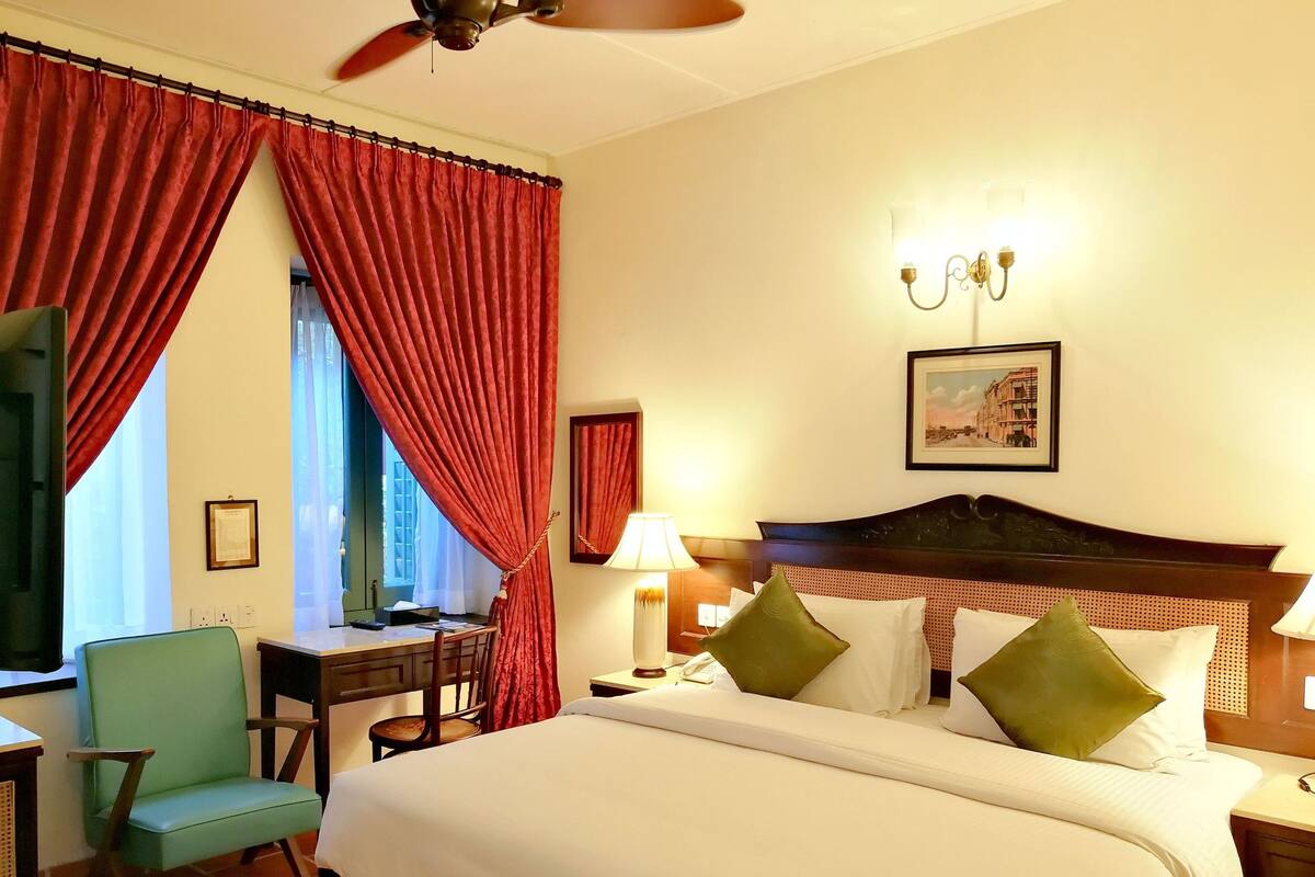 A hotel room with a bed, a desk, a chair, a window with curtains, a ceiling fan, and a wall-mounted lamp.
