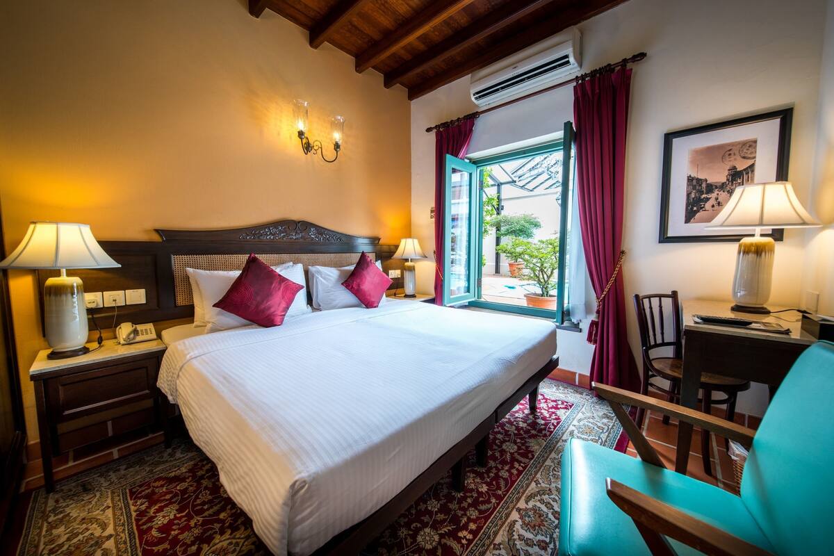 A well-appointed hotel room with a large bed, two bedside tables with lamps, a desk with a chair, and a view through an open window.