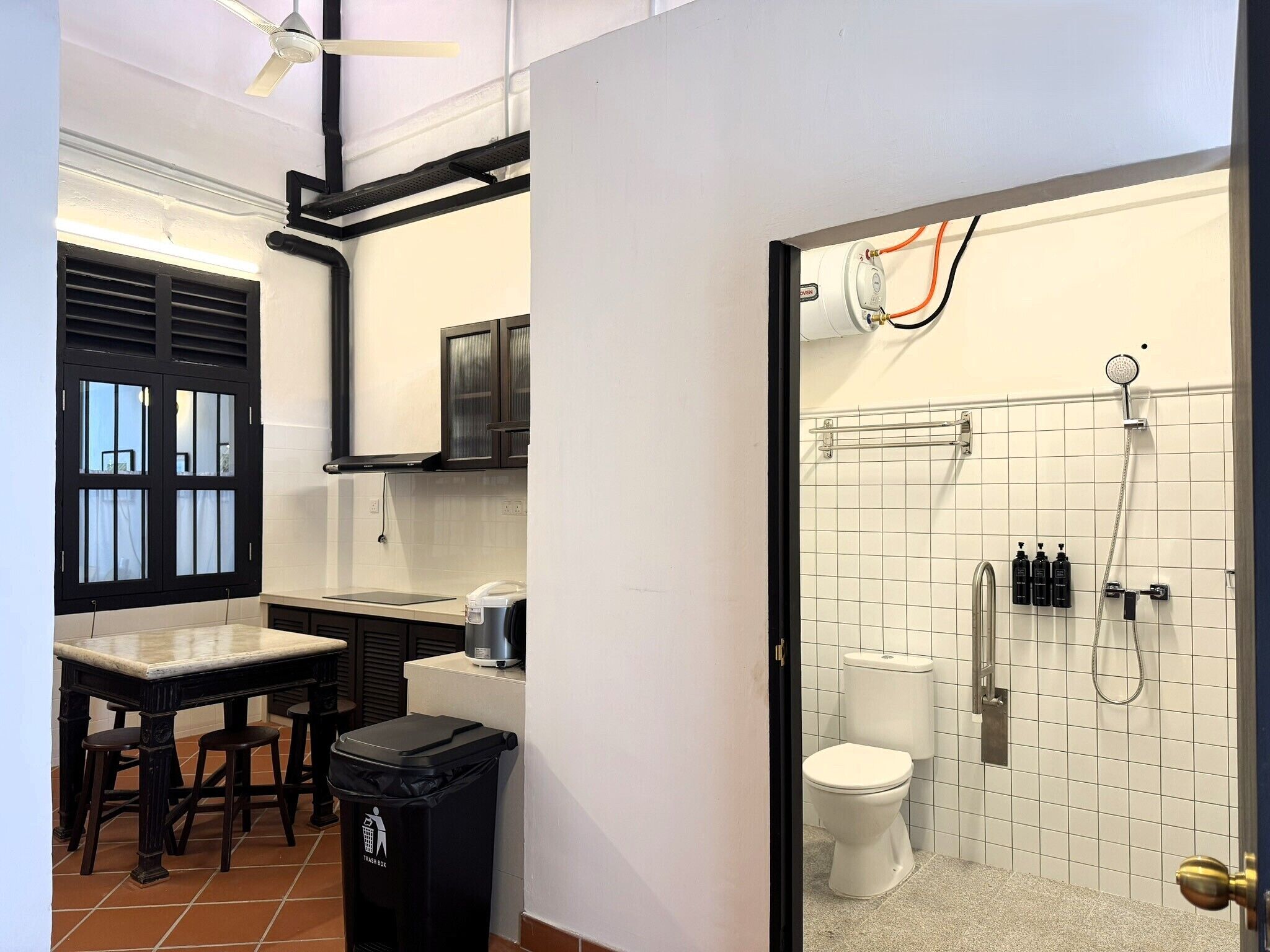 A compact kitchen with a dining area, a trash bin, and a view into a bathroom with a toilet and shower.