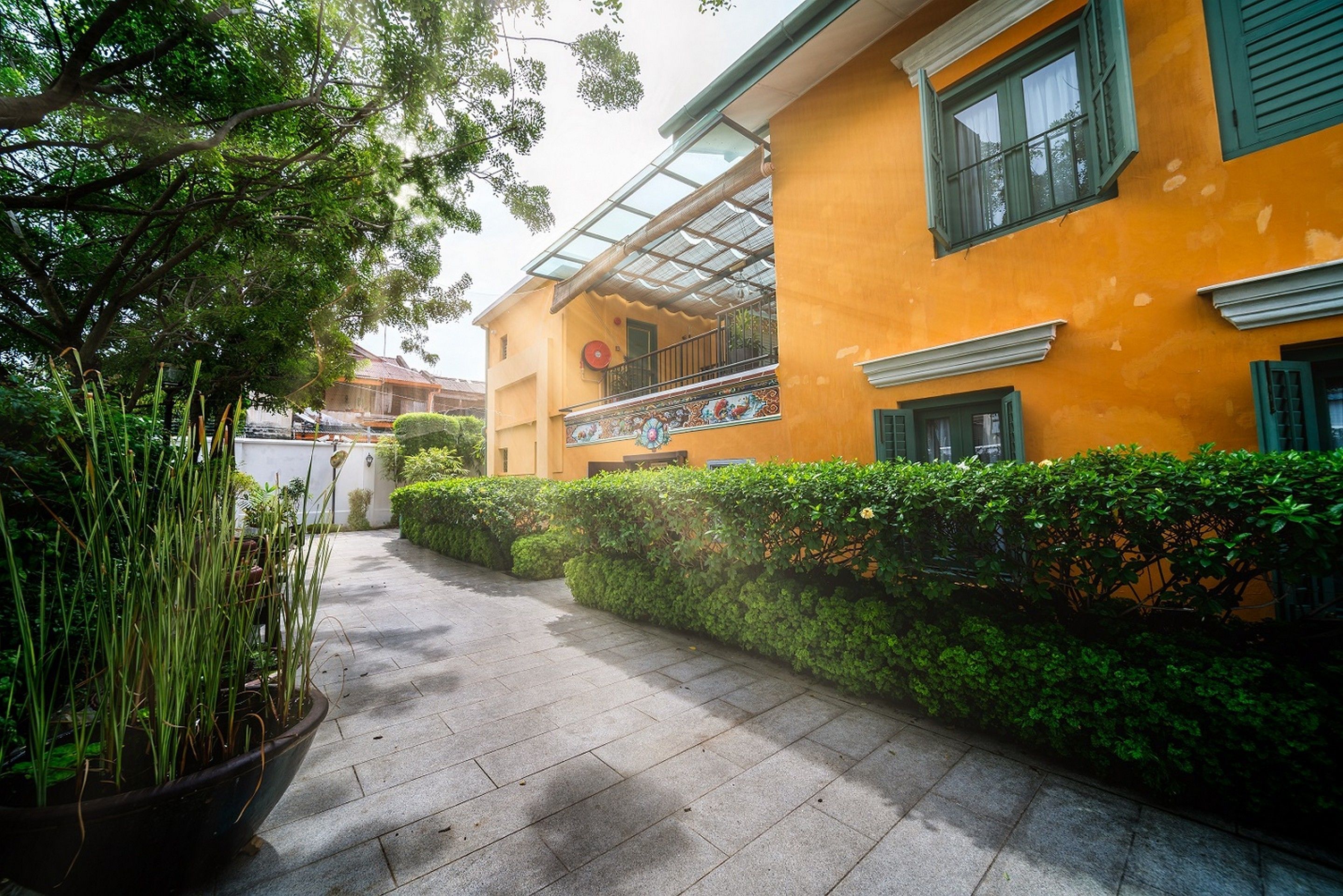 A pathway leading to a yellow building with a balcony, surrounded by greenery and potted plants.