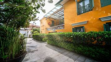 A pathway leading to a yellow building with a balcony, surrounded by greenery and potted plants.