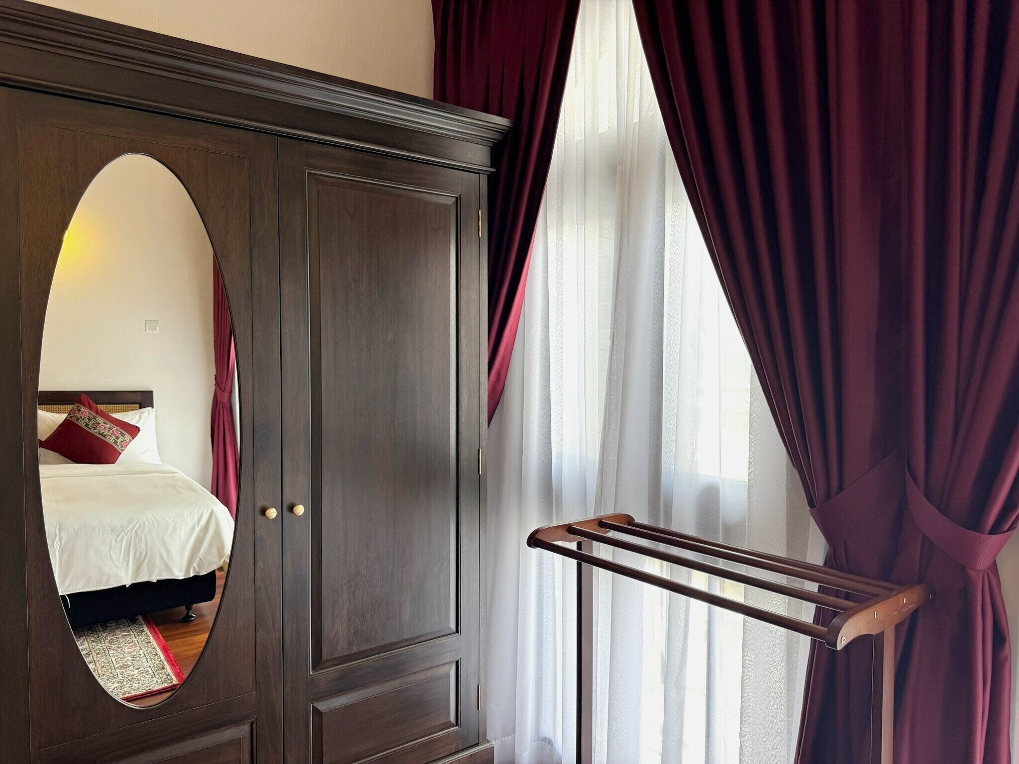 Quarto com guarda-roupa de madeira escura, um espelho, uma cama com roupa de cama branca e uma janela com cortinas finas brancas e cortinas vermelhas escuras.