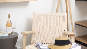 A wicker chair with a striped towel and a hat, a floor lamp, a wine bottle, and a glass on a round table. - Endless Coast, A Boutique Hotel (Wellfleet)