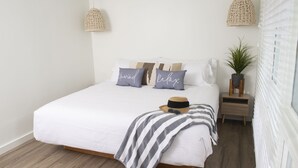 A neatly made bed with a striped blanket, two pillows with text, a hat, and a small plant on a nightstand. - Endless Coast, A Boutique Hotel (Wellfleet)