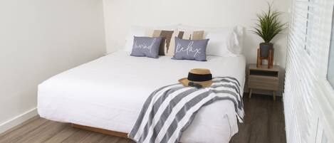A neatly made bed with a striped blanket, two pillows with text, a hat, and a small plant on a nightstand.