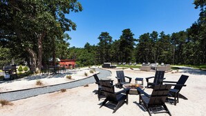 An outdoor seating area with Adirondack chairs and a fire pit, surrounded by sand and trees. - Endless Coast, A Boutique Hotel (Wellfleet)