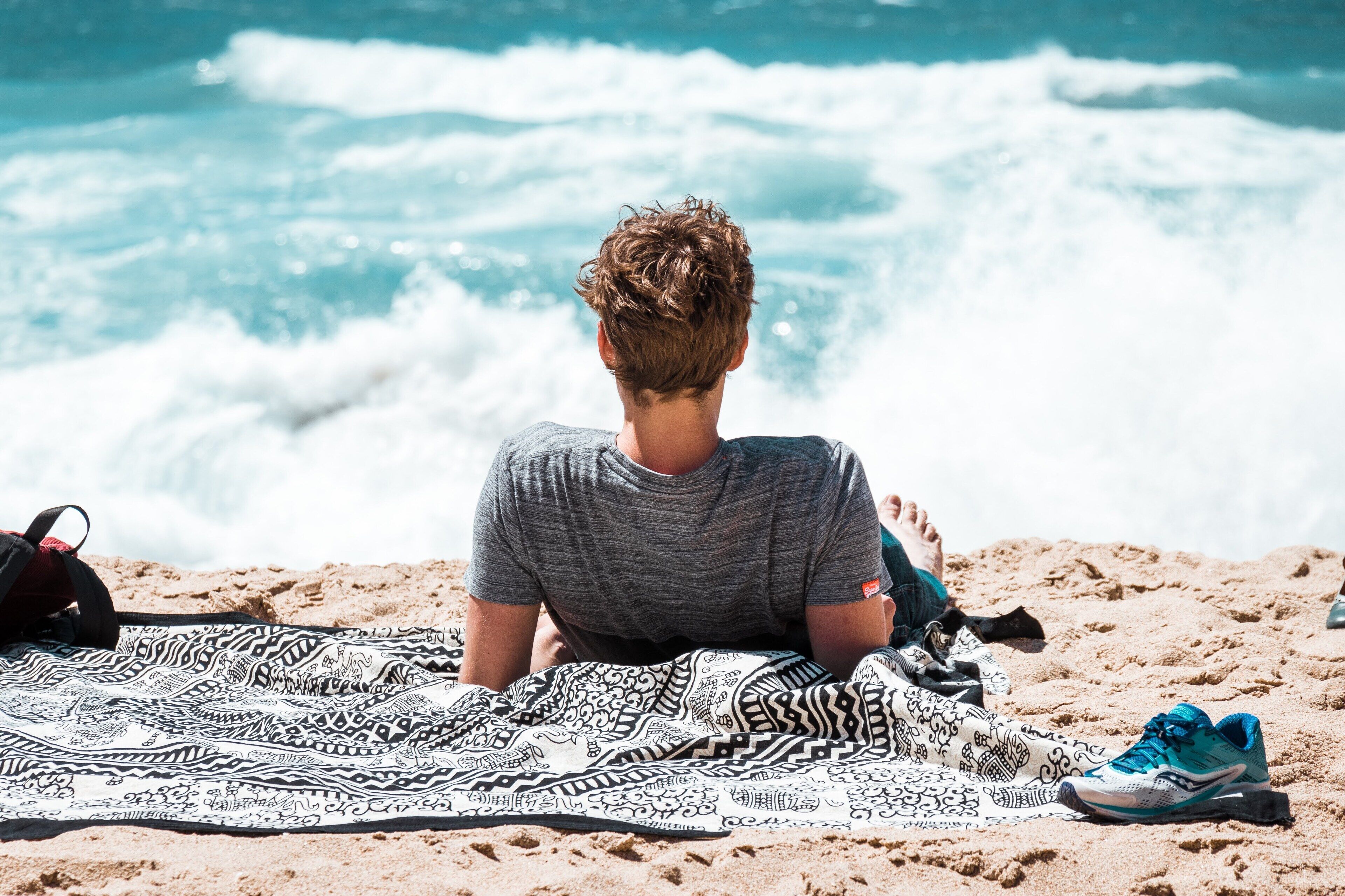 A person sitting on a beach towel facing the ocean, with a backpack and shoes nearby.