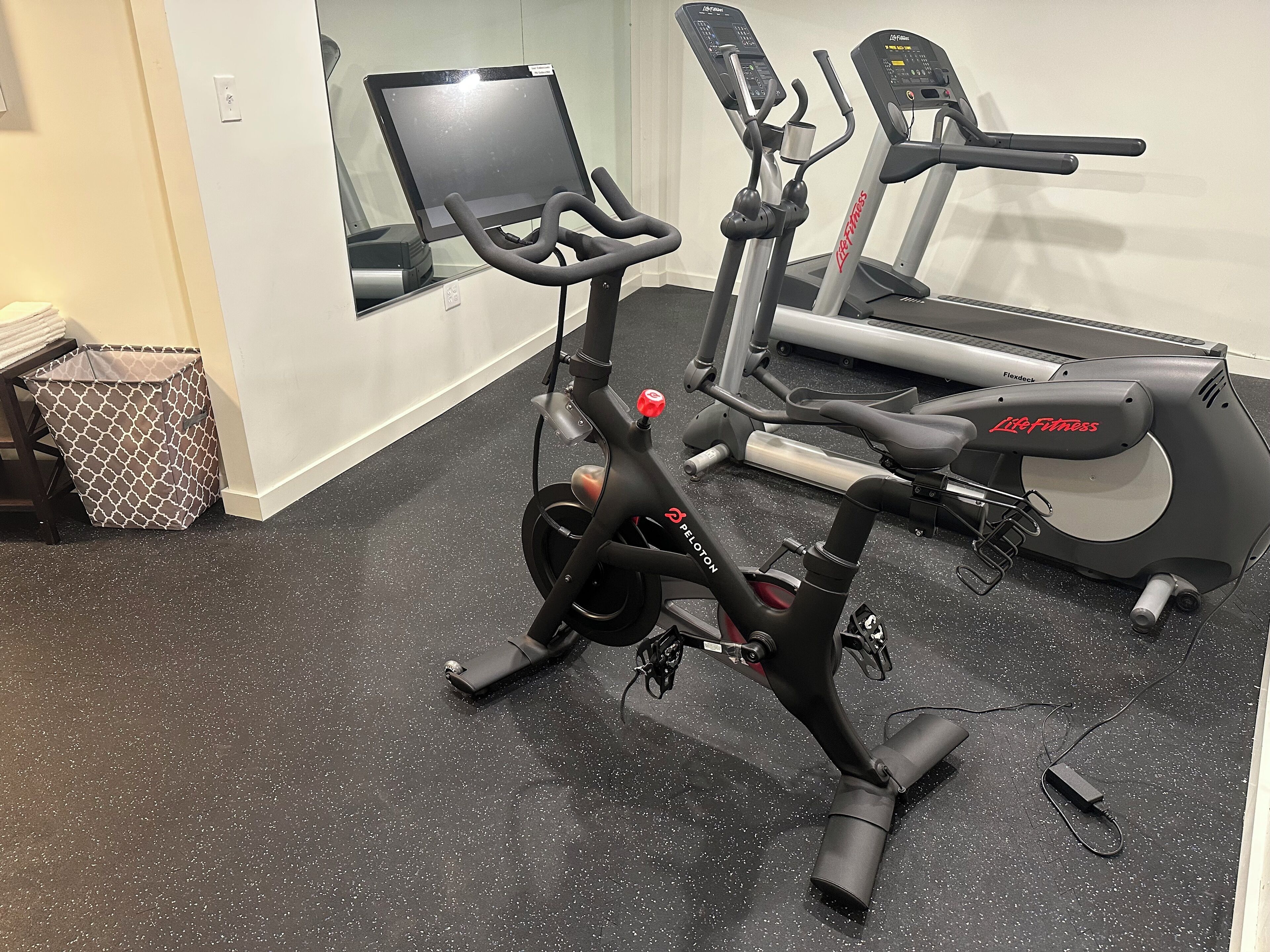 Un vélo d’appartement et un tapis de course se trouvent dans une pièce dont le sol est recouvert d’un motif moucheté, et qui contient également une poubelle et une fenêtre.
