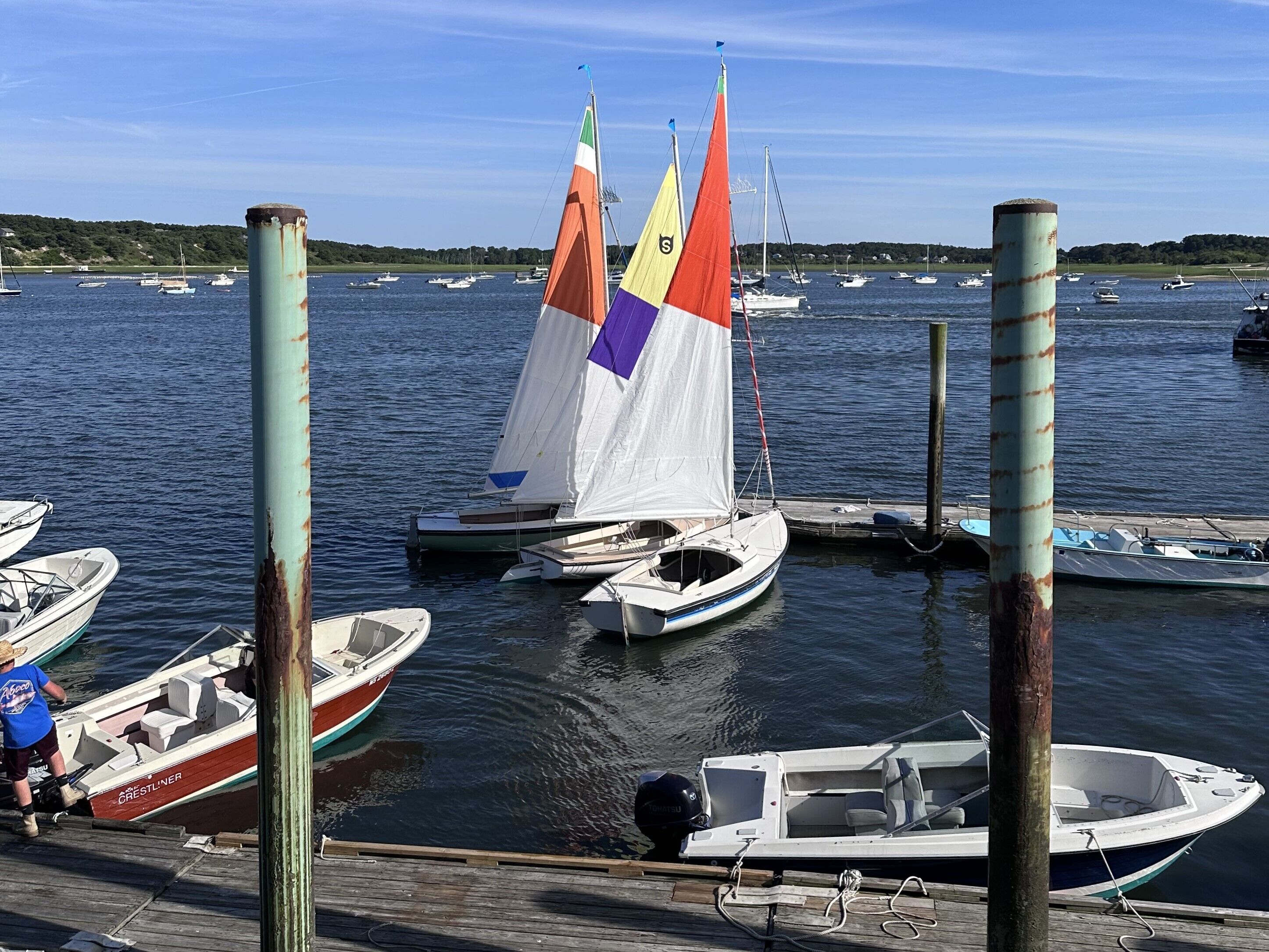 Un puerto deportivo con varios veleros amarrados, un muelle de madera y una persona con camisa azul junto a un barco.