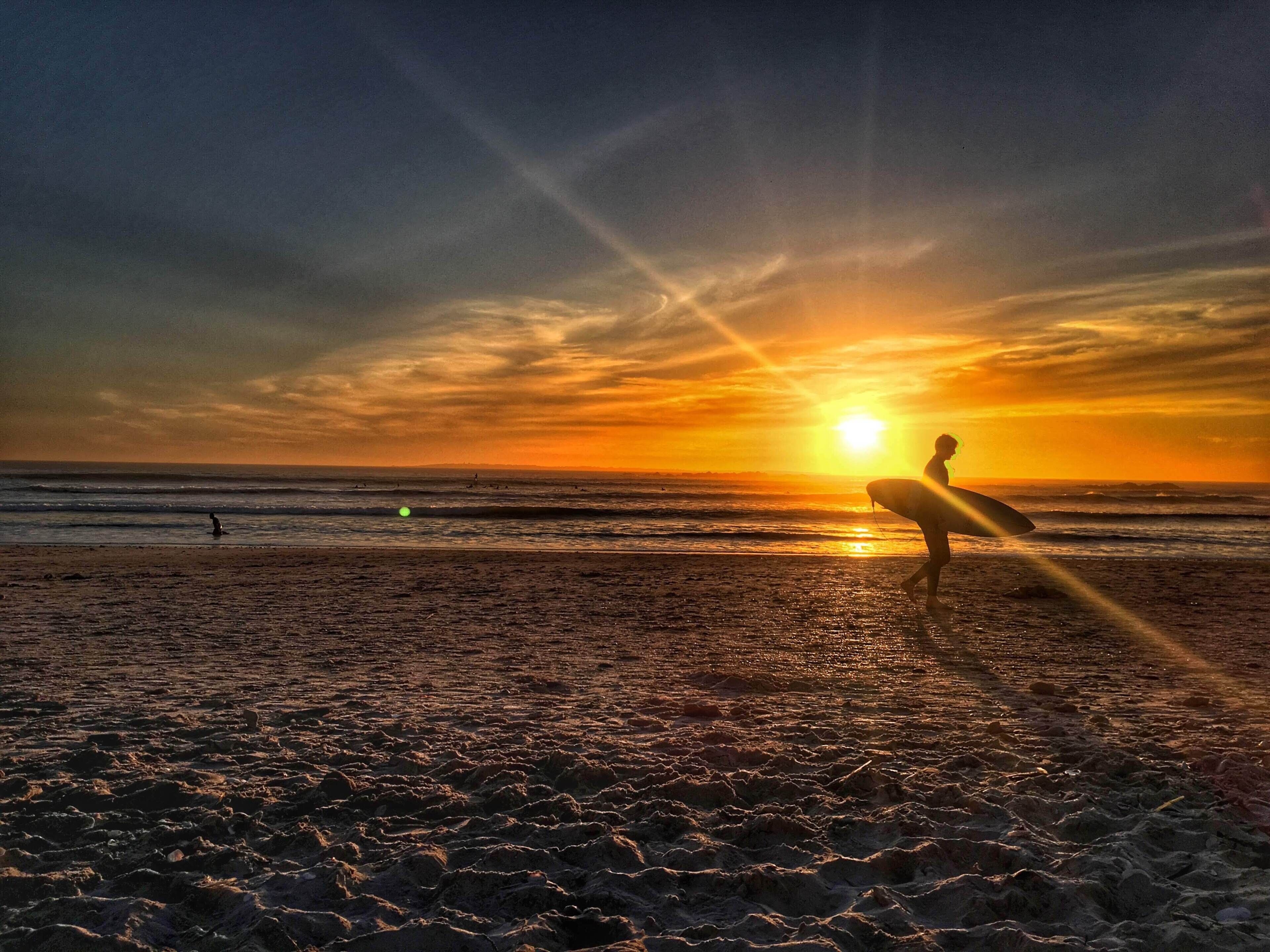 Une scène de plage avec un coucher de soleil, des vagues et une personne tenant une planche de surf.