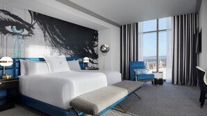 A modern hotel room with a large bed, a bench, a desk, and a view of the city.