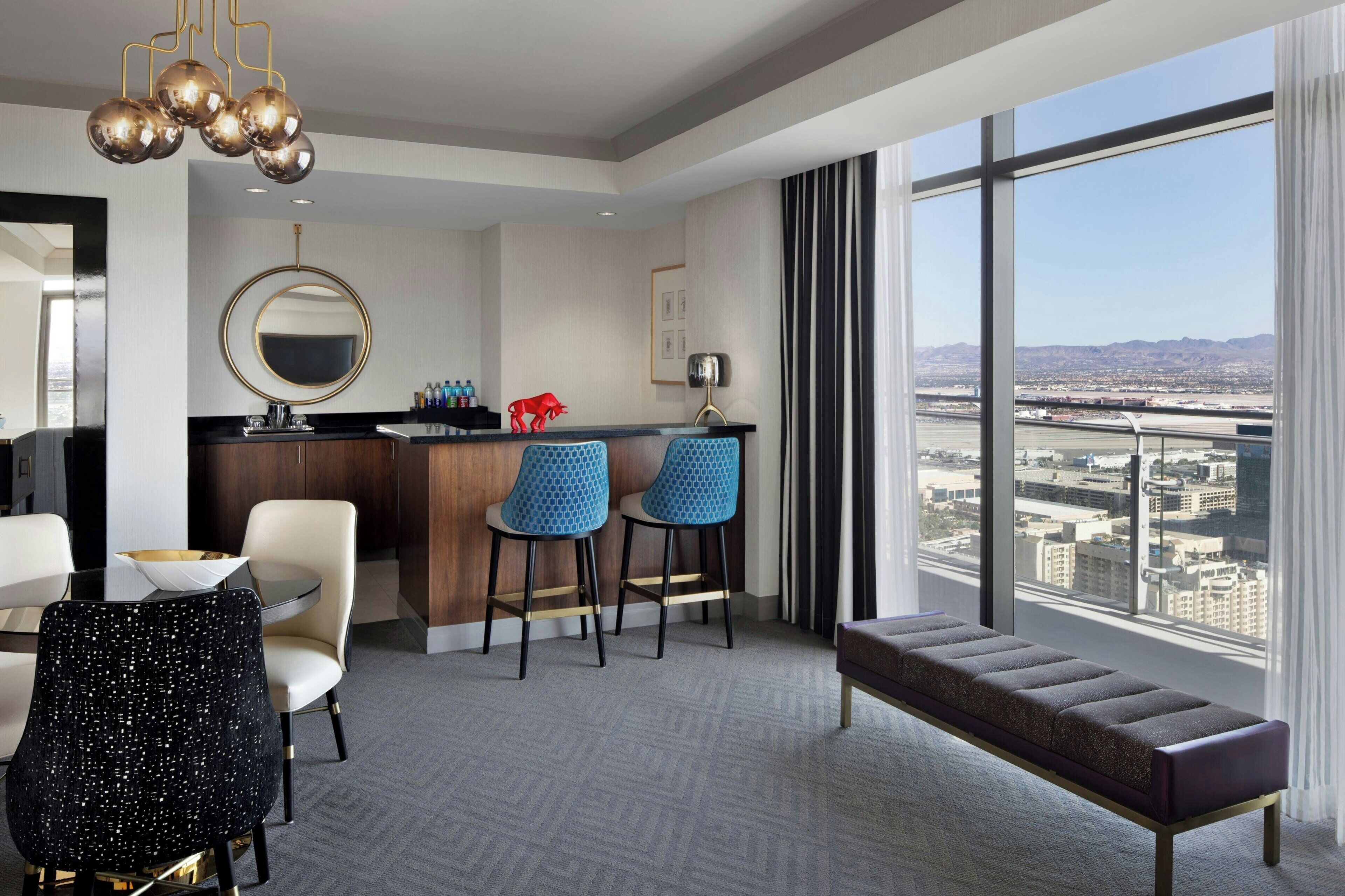 Quarto de hotel moderno com uma janela ampla que oferece vista para a cidade, um espelho redondo, área de bar com banquetas e um espaço de estar com um banco e cadeiras.