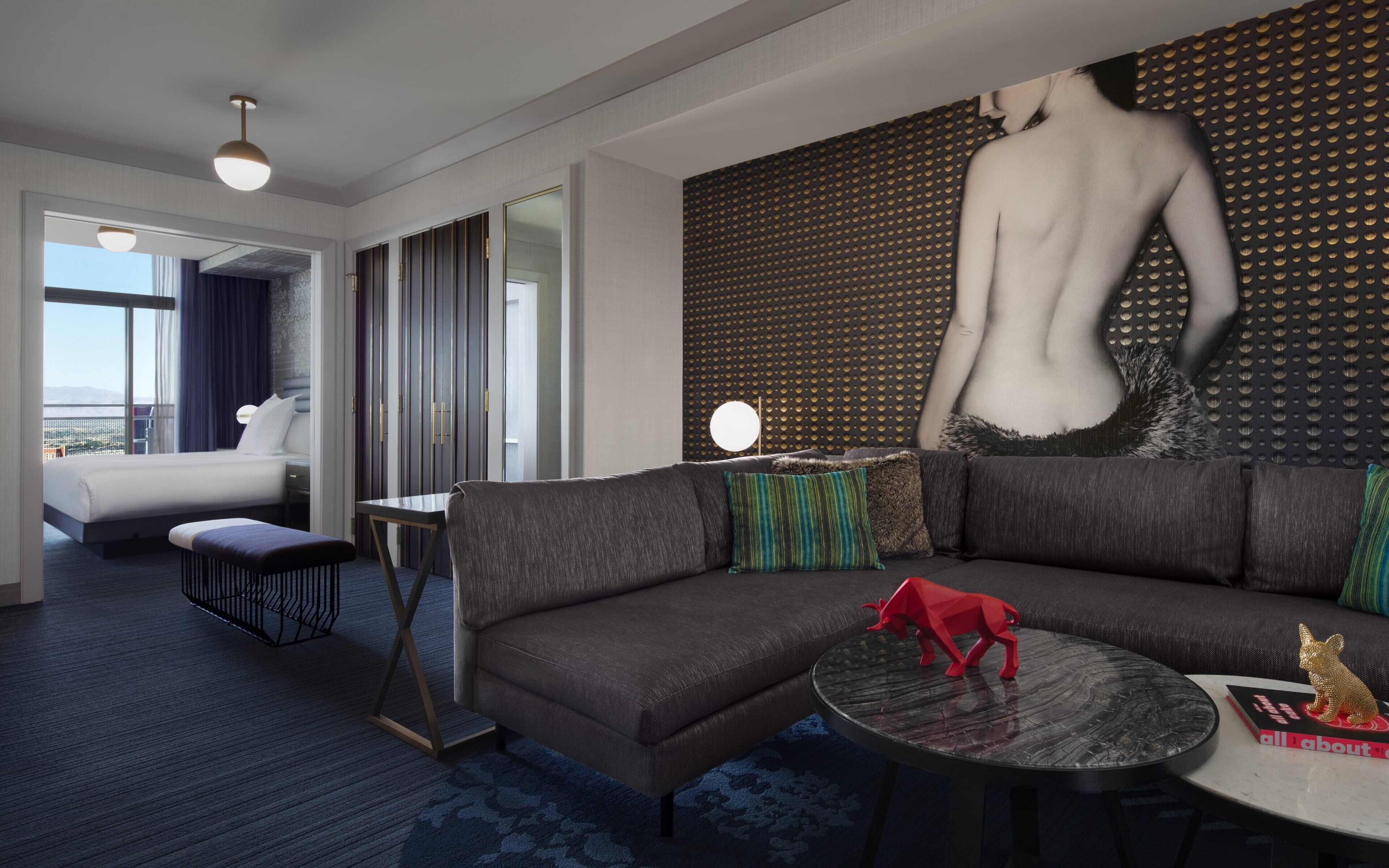 A modern hotel room with a large sectional sofa, a round coffee table, and a large painting of a nude figure. There is a bed with a view of the cityscape, a small table with a lamp, and a large window.