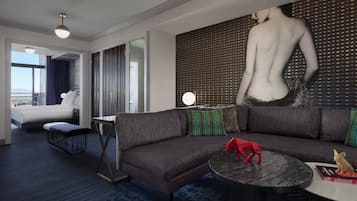 A modern hotel room with a large sectional sofa, a round coffee table, and a large painting of a nude figure. There is a bed with a view of the cityscape, a small table with a lamp, and a large window.