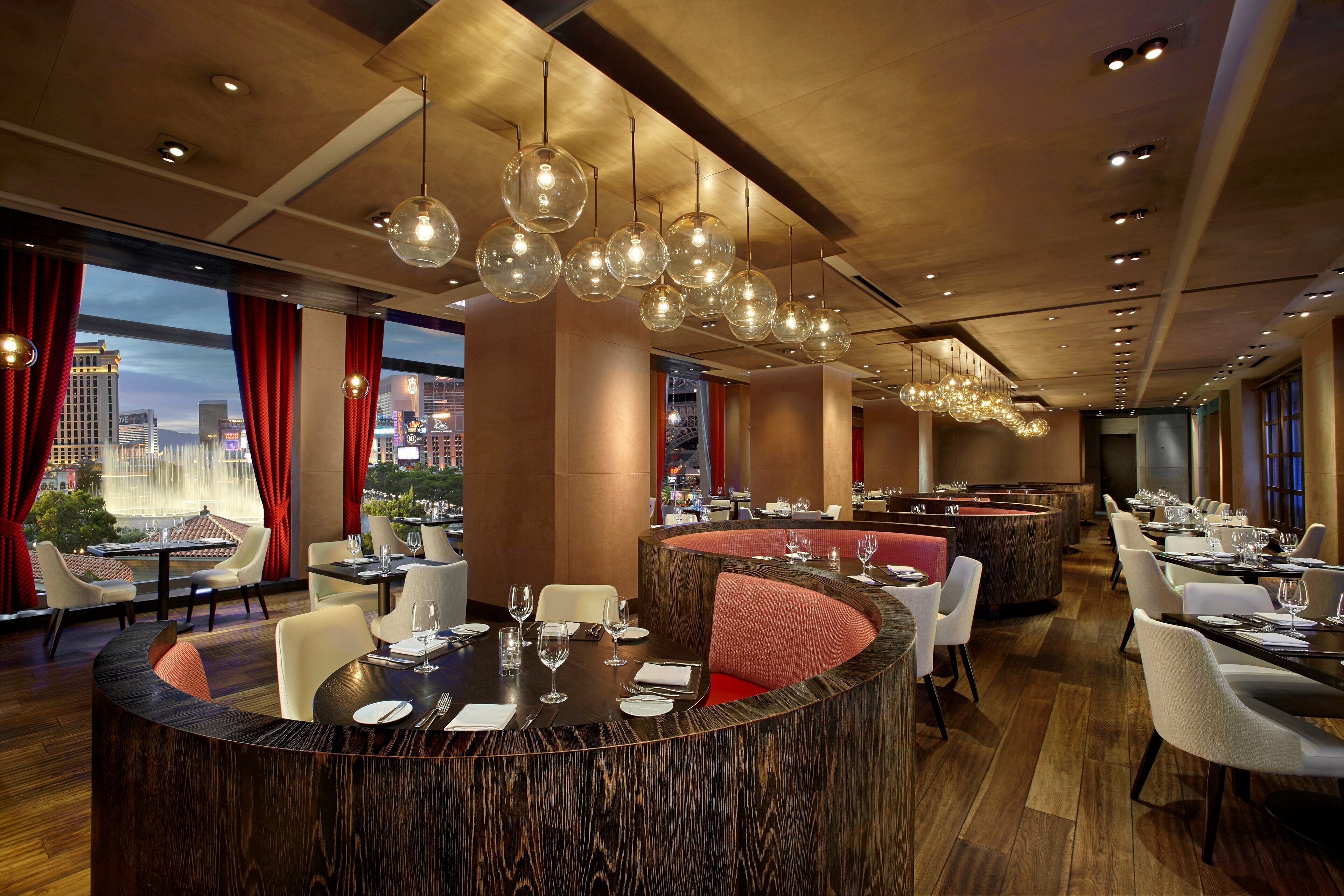 A modern restaurant with a central bar, multiple dining tables, and a city view.