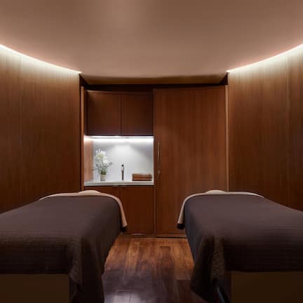 A room with two beds, wooden walls, a small window with a plant, and a shelf with items.