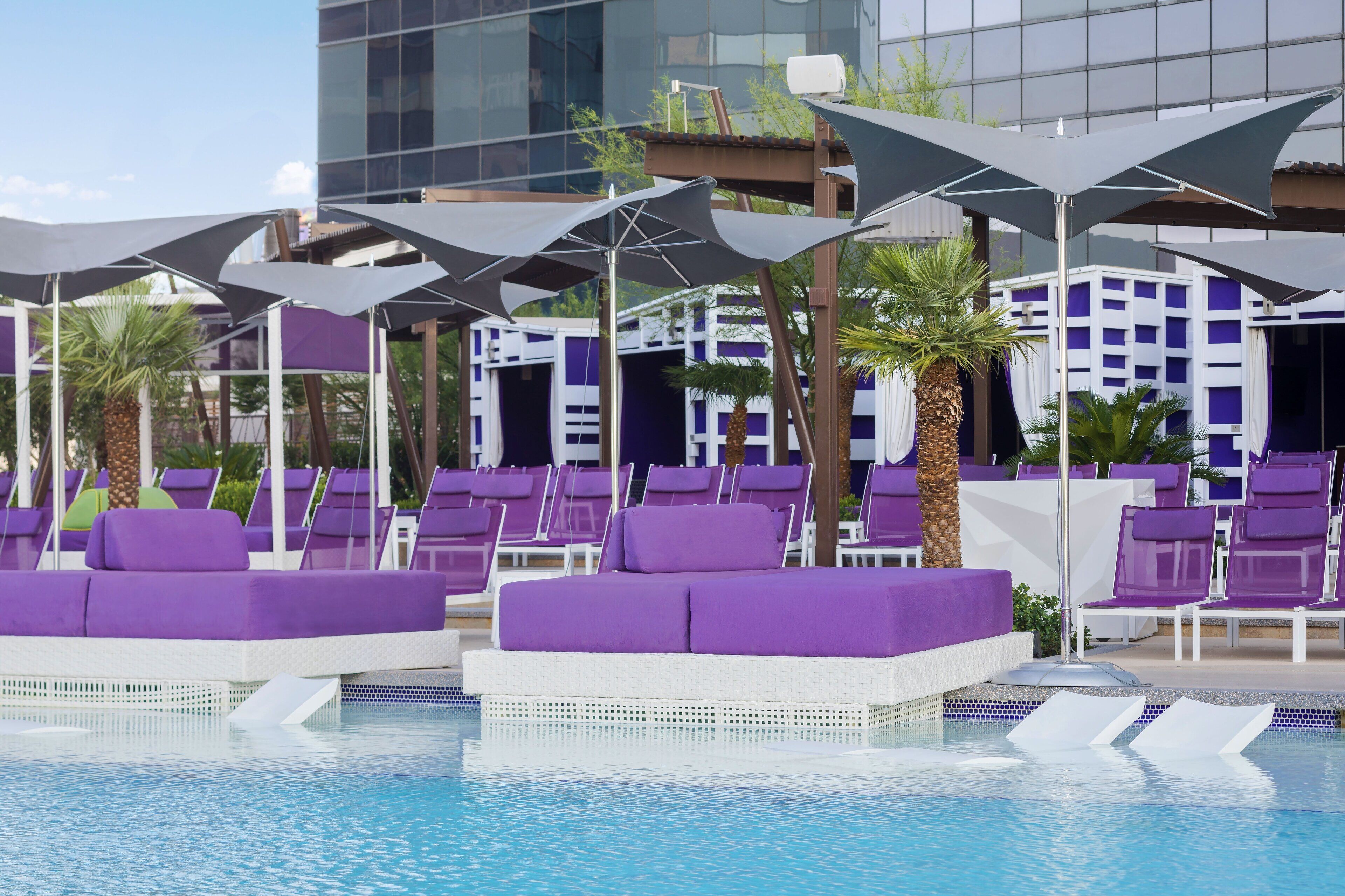 Ein Poolbereich mit violetten Liegestühlen und Sonnenschirmen, einem klaren blauen Pool und einem modernen Gebäude im Hintergrund.