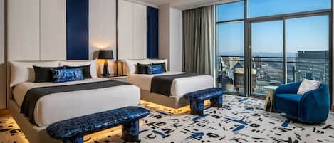 Habitación de hotel con dos camas, un ventanal grande con vista a la ciudad, un sillón azul y una alfombra estampada.