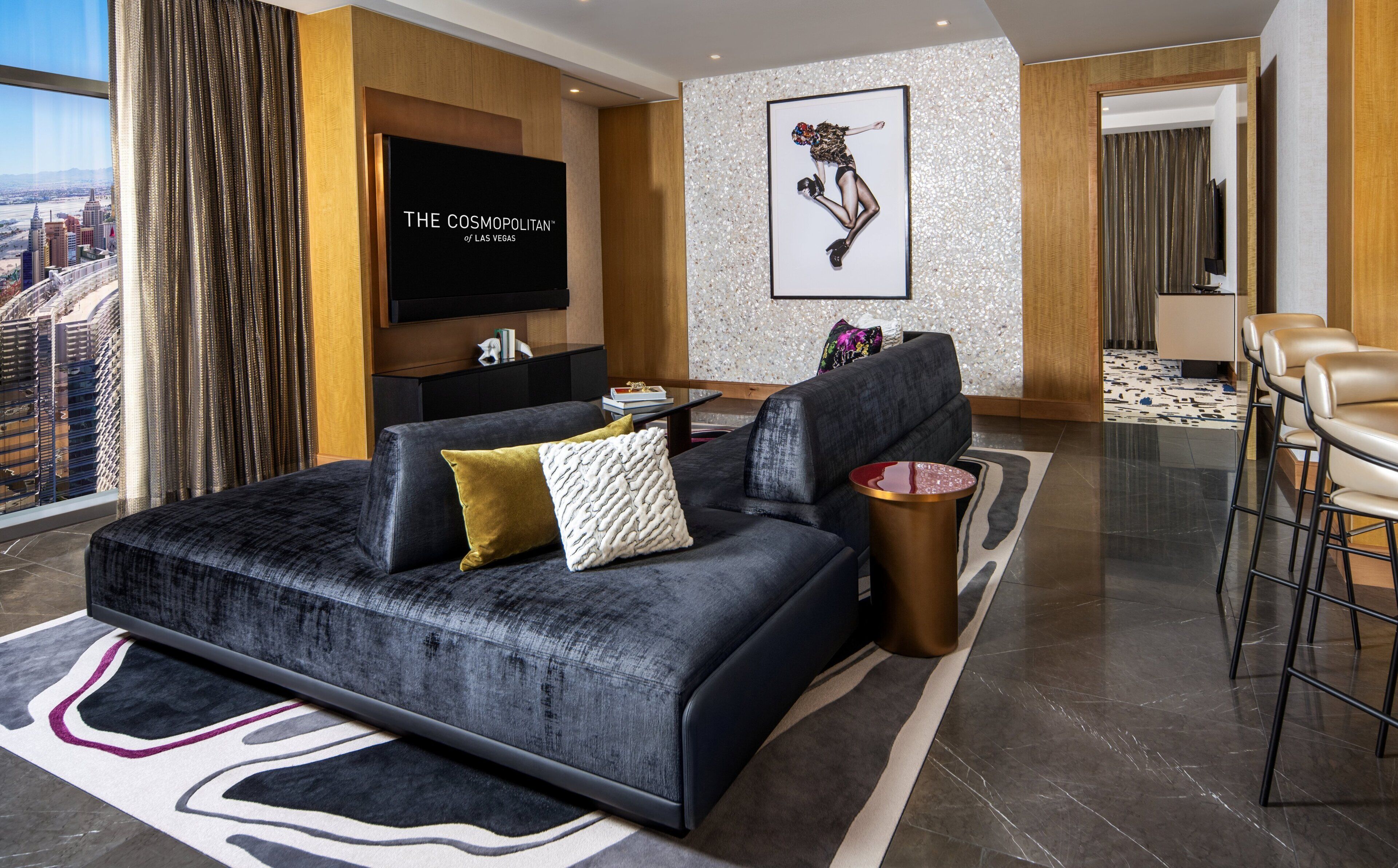 A modern living room with a large black sofa, a flat-screen TV, and a cityscape view.