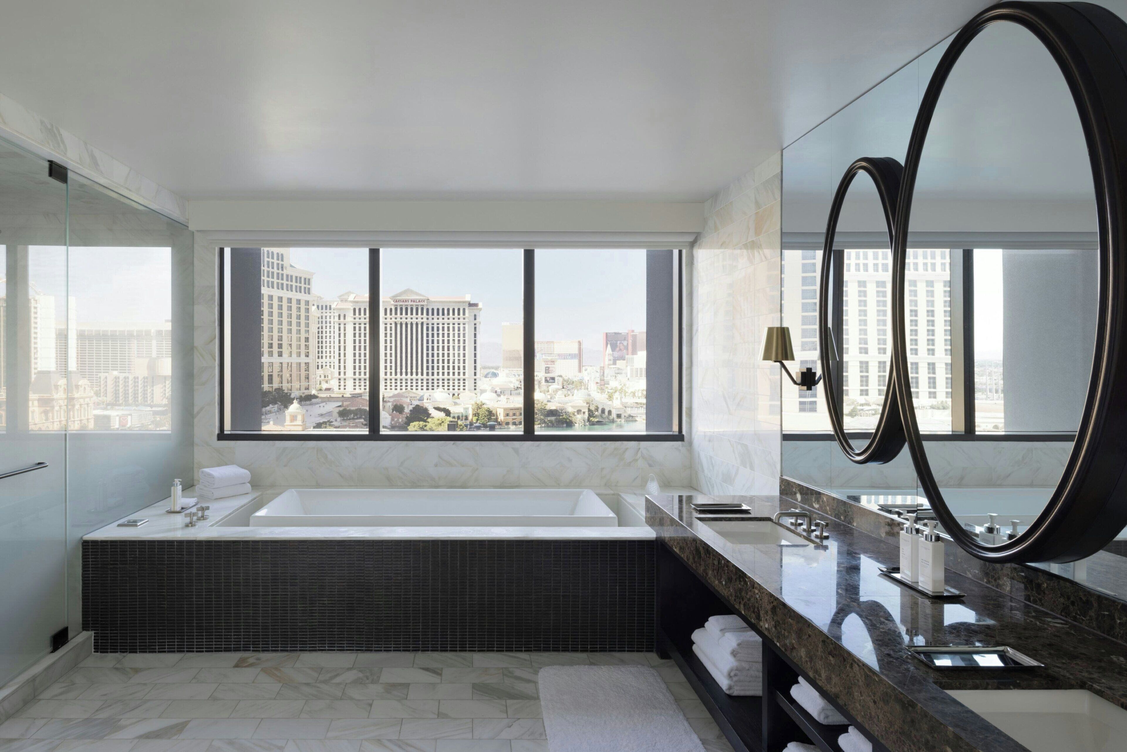 Une salle de bain moderne dotée d’une grande baignoire, d’un meuble-lavabo avec un miroir et offrant une vue sur le paysage urbain grâce à de grandes fenêtres.