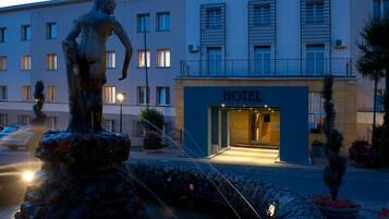 A hotel building with a statue in the foreground, a well-lit entrance, and a paved area with potted plants.