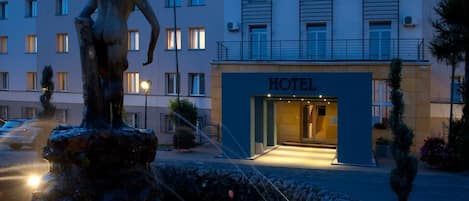 A hotel building with a statue in the foreground, a well-lit entrance, and a paved area with potted plants.