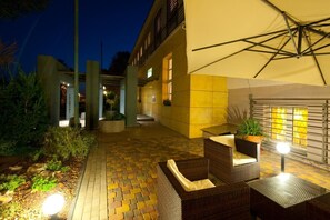 An outdoor patio area with wicker furniture and a yellow umbrella, surrounded by a brick pathway and greenery.