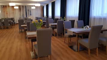 A hotel dining area with multiple tables and chairs, large windows with curtains, and hanging light fixtures.