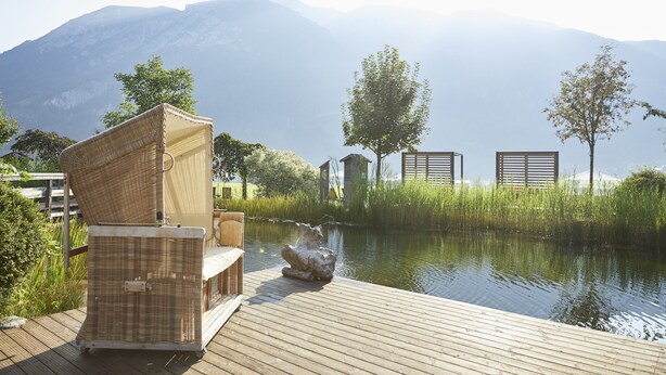 A wooden deck with wicker furniture, a stone sculpture, and a view of a calm lake with mountains in the background. - Familienparadies Sporthotel Achensee - All Inclusive (Achenkirch)