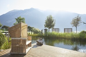 A wooden deck with wicker furniture, a stone sculpture, and a view of a calm lake with mountains in the background. - Familienparadies Sporthotel Achensee - All Inclusive (Achenkirch)