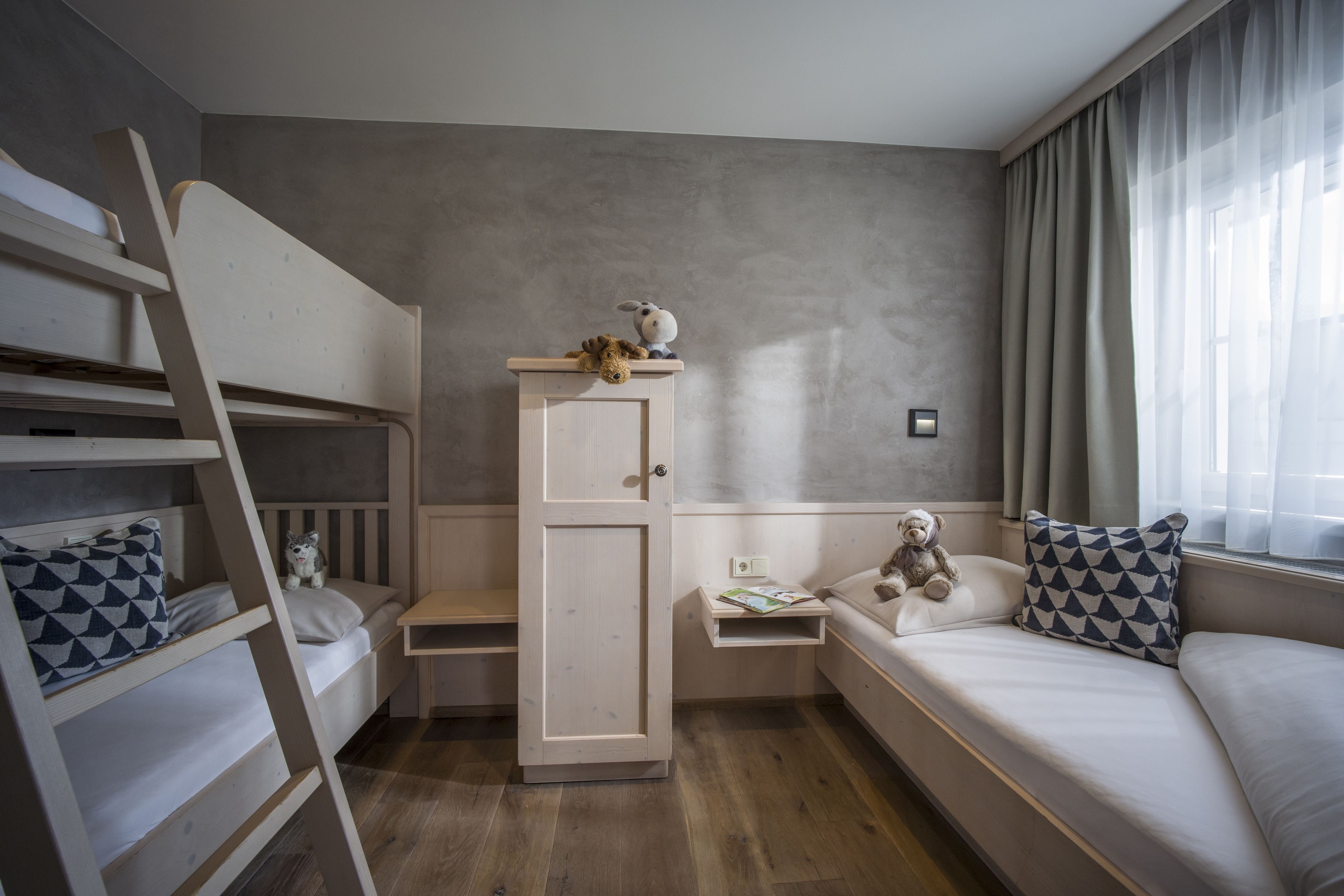A bedroom with bunk beds, a wardrobe, a bedside table with a teddy bear, and a window with curtains.