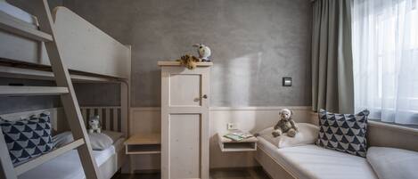 A bedroom with bunk beds, a wardrobe, a bedside table with a teddy bear, and a window with curtains.