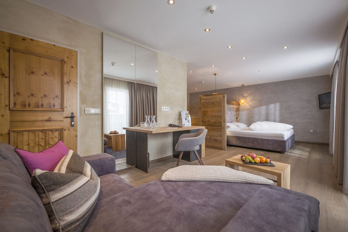 A modern hotel room with a sofa, a small desk, a bed, and a dining table with fruit.