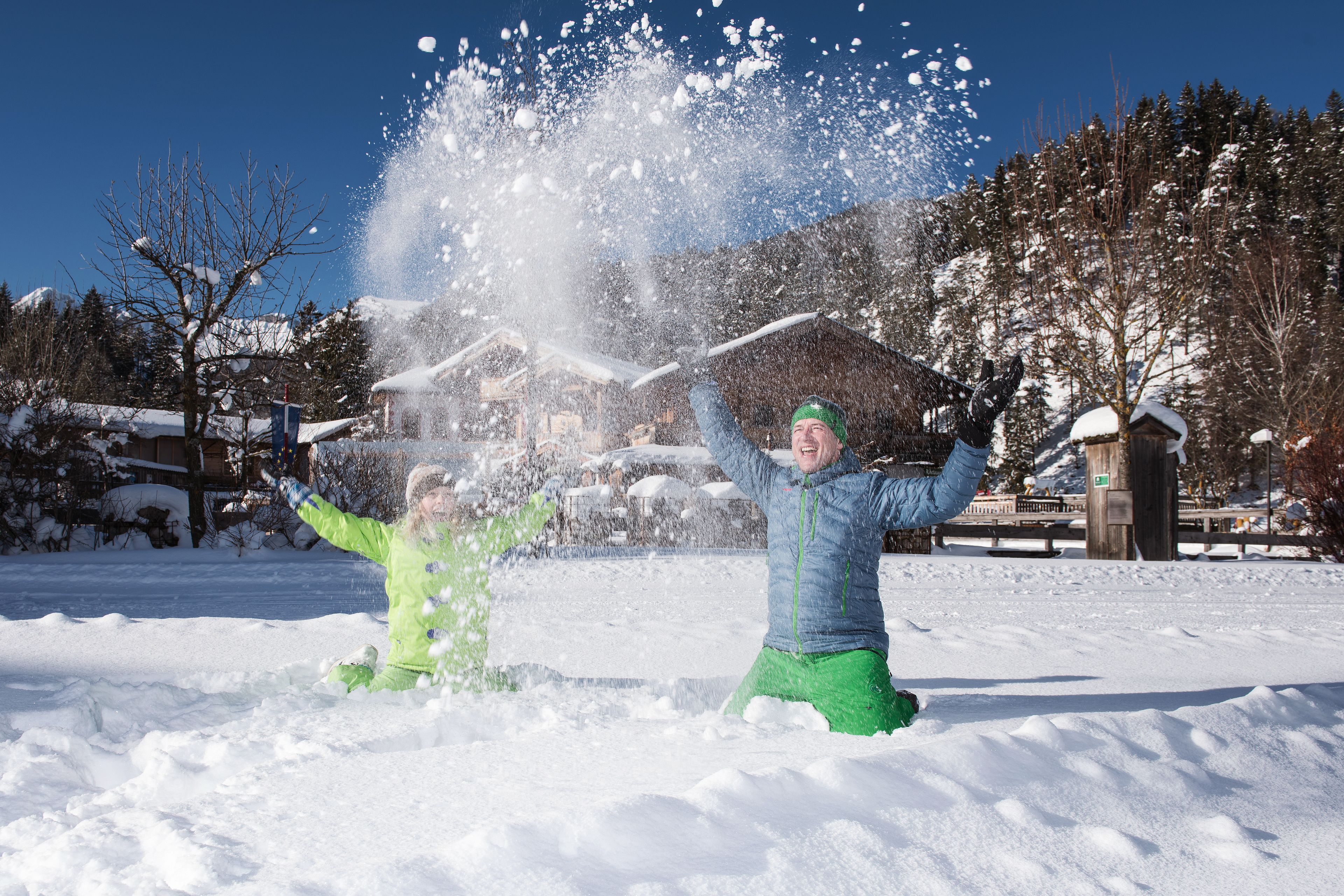 Escena nevada al aire libre con una persona con chaqueta azul y pantalones verdes lanzando nieve.