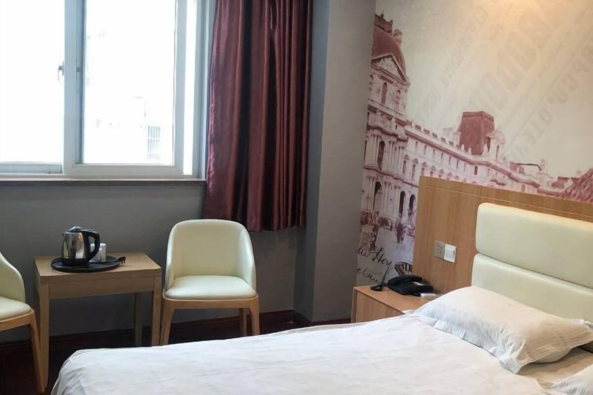 Habitación de hotel con cama, escritorio con hervidor, dos sillas, mesita de noche y cortina.