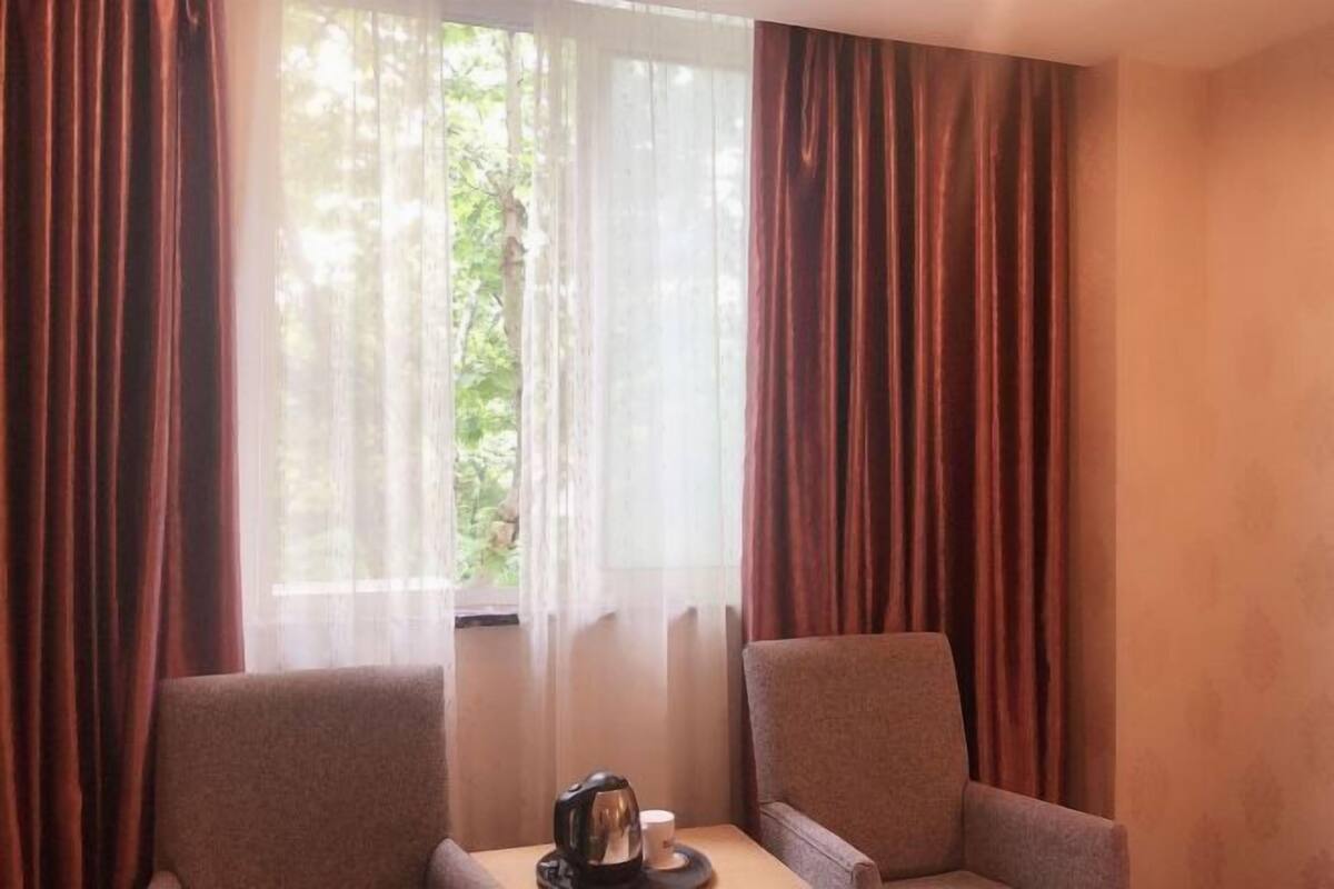 Una habitación con dos sillones, una mesa con cafetera y taza, y cortinas ligeras sobre una ventana.